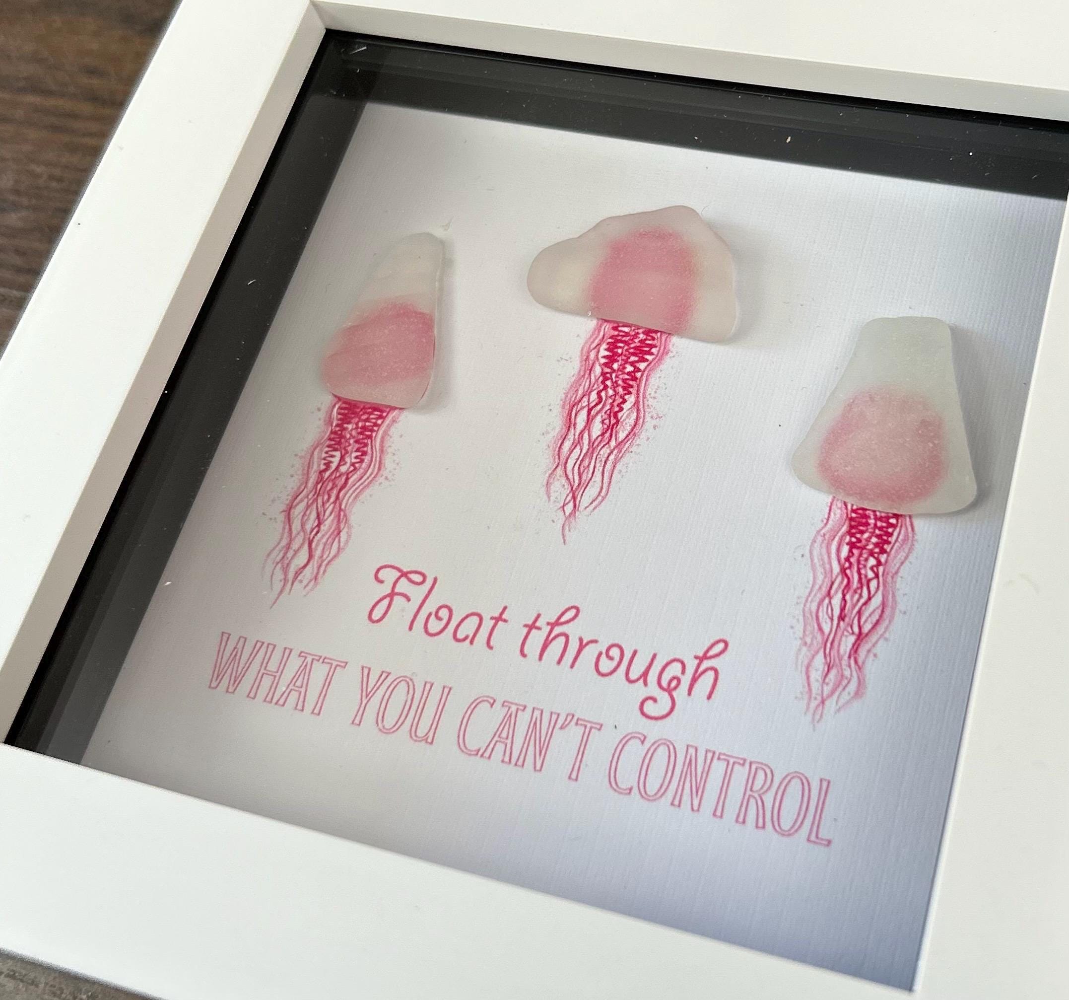 Pink sea glass jellyfish art in white frame with calming coastal quote wall decor in a home setting