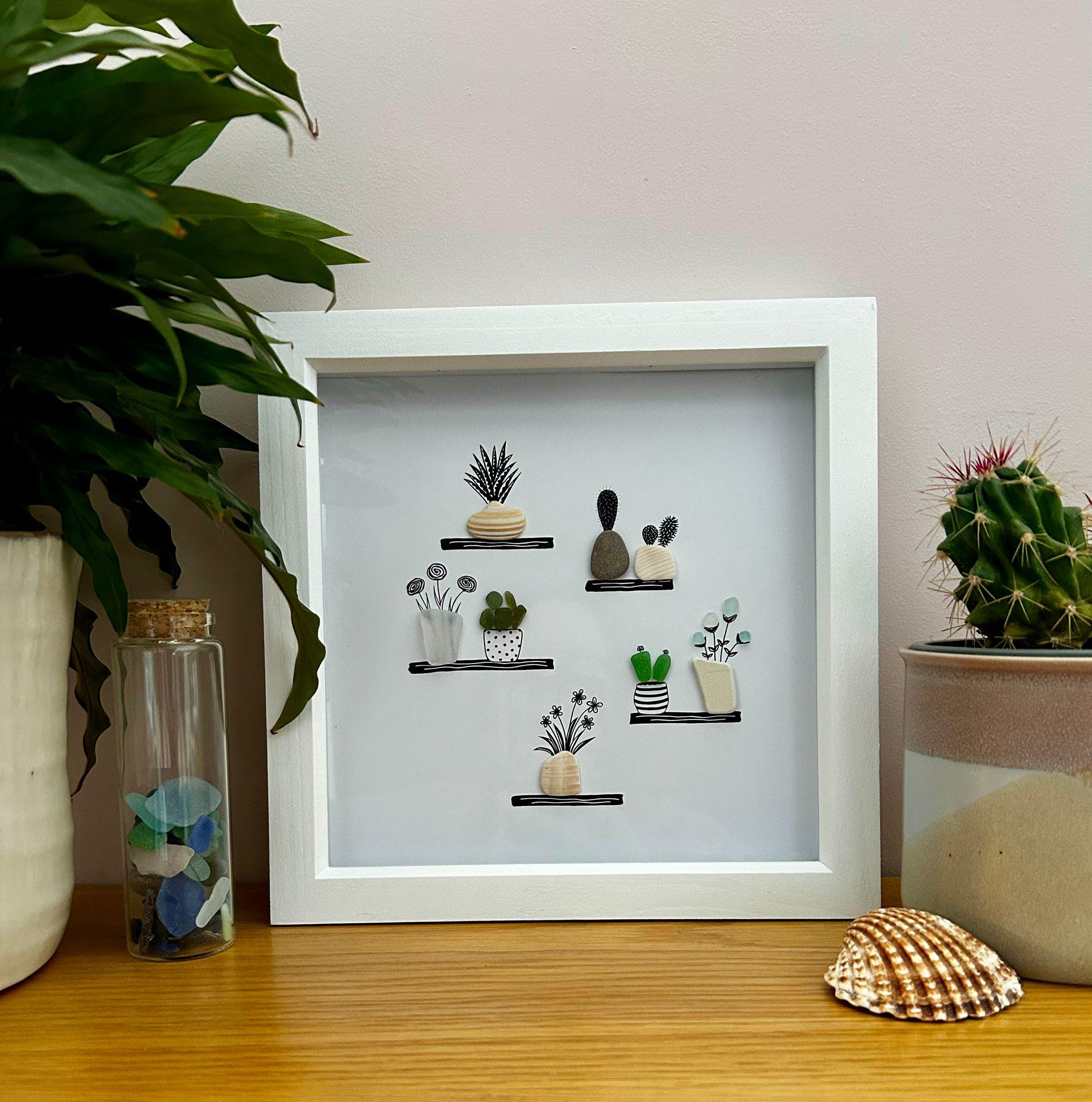 May include: A white framed artwork featuring pebble art plants on black shelves. The artwork includes various potted plants, such as cacti and flowers. A glass bottle with sea glass is in front of the artwork.